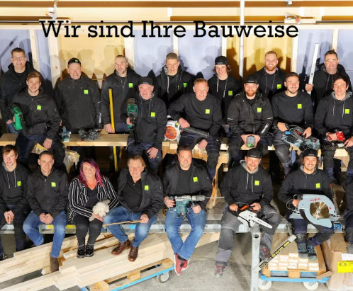 Ein Team von Handwerkern in Arbeitskleidung posiert in einer Werkstatt mit Werkzeugen und Baumaterialien. Text: Wir sind Ihre Bauweise.