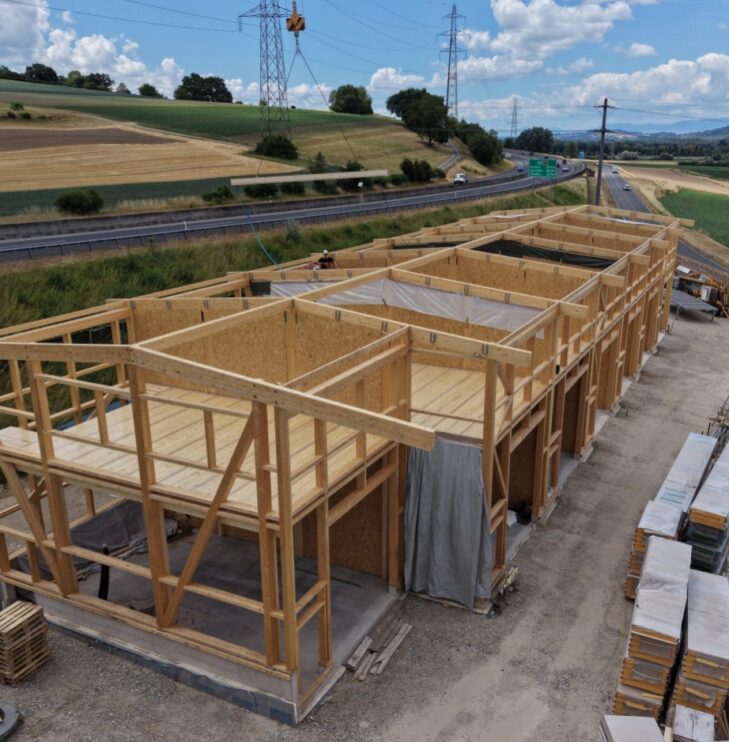 Holzrahmenbau eines Gebäudes neben einer Landstraße unter blauem Himmel mit Wolken im Hintergrund.