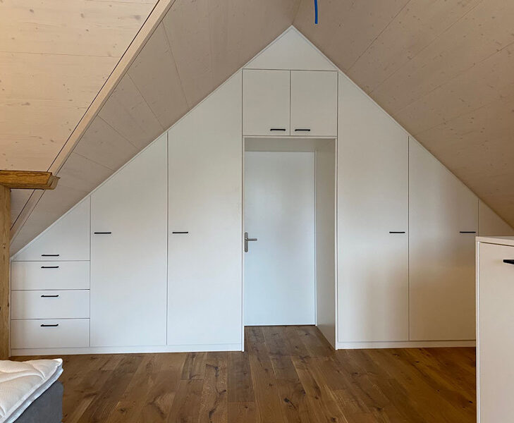 Einbauschrank in Dachschräge aus weißem Holz mit mehreren Türen und Schubladen, vor Holzboden.