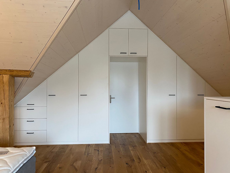 Einbauschrank in Dachschräge aus weißem Holz mit mehreren Türen und Schubladen, vor Holzboden.