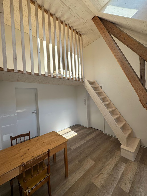 Heller Raum mit Holztreppe, Zwischengeschoss, Tisch und Stuhl, Dachschräge und Holzbalken im modernen, minimalistischen Design.