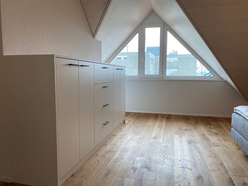 Modernes Dachgeschosszimmer mit Holzfußboden, weißem Einbauschrank und großem Fenster für viel Tageslicht.