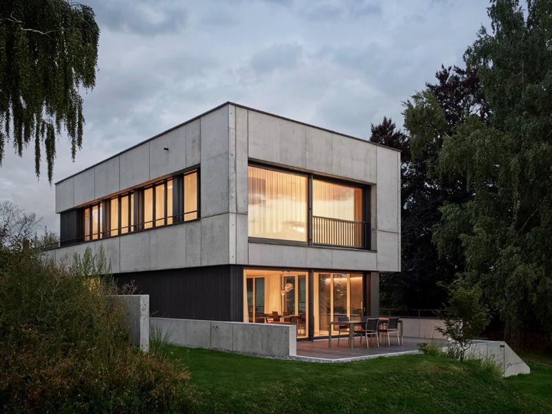 Modernes zweistöckiges Haus aus Beton mit großen Fenstern und Garten bei Sonnenuntergang.