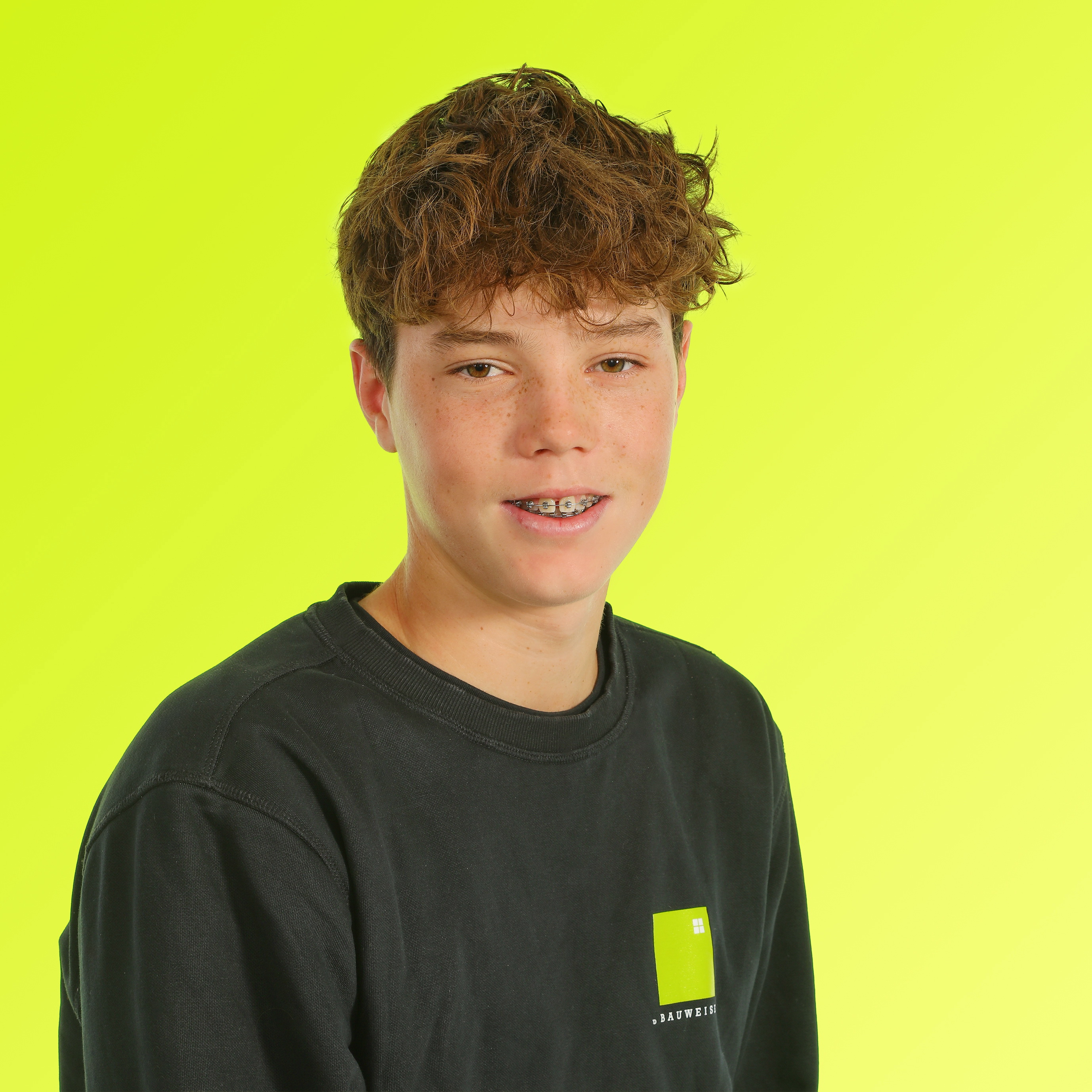 Junger Mann in schwarzem Shirt vor gelbem Hintergrund, lockiges Haar, freudiger Ausdruck, Zahnspange sichtbar.